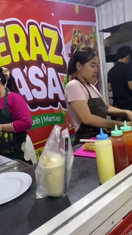 The video shows two women preparing food at a food stall with a red and yellow sign that says "TERAZ ASA". The woman on the left is wearing a pink shirt and a green apron and appears to be cooking something in a pan. The woman on the right is wearing a pink shirt and a brown apron and is wrapping something in paper. In front of them are various food items, condiments in bottles, and a drink in a plastic cup. The environment appears to be outdoors or in a semi-enclosed space, possibly a market or street food area.