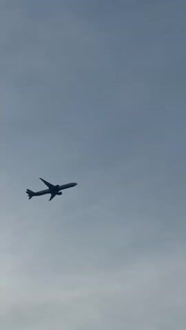 An airplane is silhouetted against a pale blue sky with scattered clouds. The plane is in mid-flight, with its wings spread and engines visible. The camera is angled upwards, capturing the plane as it flies across the frame. The lighting suggests it is daytime, possibly late afternoon, given the warm tones in the clouds towards the end of the clip. There are no people visible in the video. The environment is entirely the sky.