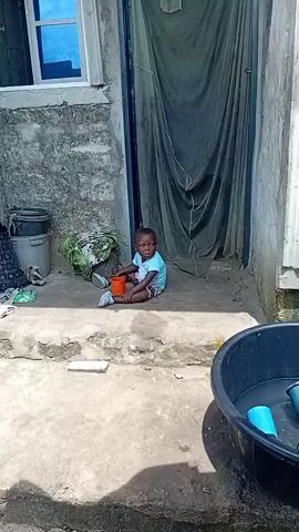 A young child sits on the ground in front of a doorway. The child is wearing a blue t-shirt and patterned shorts. A red cup is to the child's left. A green and white patterned cloth is behind the child. To the right of the child, a black basin holds several blue cups. The environment appears to be outdoors.
