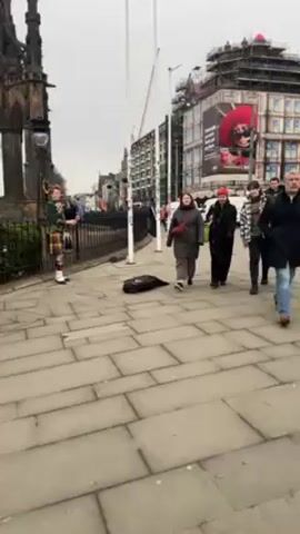 The video captures a street scene in Edinburgh, Scotland, featuring a bagpiper performing on the sidewalk. The bagpiper, dressed in traditional Scottish attire including a kilt, stands near a black, ornate structure. Pedestrians walk by on the cobblestone pavement. Among them are three young women who are walking closely together and are near the camera. Other pedestrians walk further down the sidewalk in the distance. A large building, partially covered with scaffolding and featuring a large advertisement, is visible in the background. The sky is overcast.