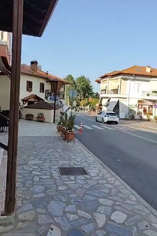 The video pans across a street scene in what appears to be a European town. On the left, a building with a covered walkway features stone paving in the foreground. Across the street, several buildings line the road. A white car is seen driving down the street, crossing a pedestrian crossing. Further down the road, a person on a scooter is seen riding away from the camera. The sky is clear and blue, and the day appears to be sunny. There are trees and foliage visible in the background, suggesting a green and somewhat rural setting within the town. Orange traffic cones are placed near the pedestrian crossing.