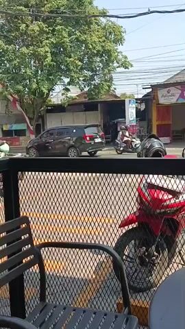 The video shows a street scene from behind a black metal fence. In the foreground, a black chair is visible. Beyond the fence, a red motorcycle is parked. The street has yellow markings on the asphalt and is bordered by buildings and trees. Several people are on motorcycles traveling on the street. One motorcycle carries a rider and a passenger. Another motorcycle has a single rider wearing a blue shirt and red helmet. A truck with a flatbed is also visible on the road. Traffic lights and electrical wires are present overhead. The sky is clear and blue.
