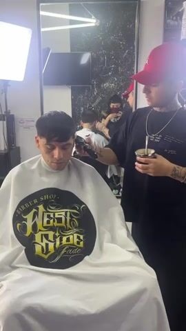 The scene is set in a barber shop. A barber with multiple tattoos, wearing a black t-shirt and a red baseball cap, is trimming the hair of a seated client with a hair clipper. The client is wearing a white barber’s cape with a stylized logo that reads “Barber Shop West Side Fade.” The barber is holding a cup, which could be a mate, in his left hand, while operating the clipper with his right. A large rectangular mirror in the background shows a reflection of the barber, the client, and another person in the background holding a phone, possibly taking a picture. Additional light is provided by a light panel to the left of the camera. Behind the mirror, there is a poster on the wall. The setting suggests a professional grooming environment.