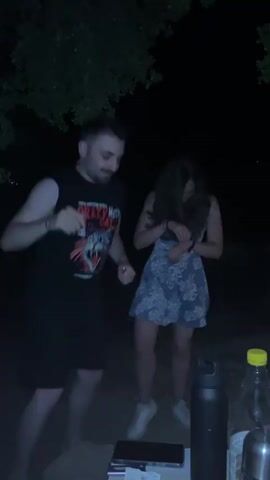 Three people are dancing outdoors at night. On the left, a man with a beard, wearing a black sleeveless shirt and shorts, moves his arms and body. In the center, a woman with long dark hair, wearing a light-colored patterned dress, is also dancing. A second woman, wearing a dark sleeveless jumpsuit or dress, enters the frame from the right, raising her right arm and shaking a small, light-colored object. A table with a black water bottle and a yellow-capped bottle is visible in the right foreground. Trees are discernible in the dark background.