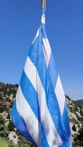 The video opens with a close-up of a Greek flag rippling in the wind against a bright blue sky. The camera then pans to reveal a mountainous landscape covered in lush green trees. A rustic wooden fence runs along a rocky path in the foreground. In the distance, the peak of the mountain is dotted with several communication towers. As the camera continues to pan, a man is seen standing near a small wooden gazebo, looking through a telescope mounted on a stand. Further to the right, a building with a stone facade and a red-tiled roof is visible. The video pans further, showing more of the building and the surrounding rocky terrain, with the wooden fence continuing to line the path. The overall scene depicts a scenic mountain viewpoint with a visitor enjoying the scenery.