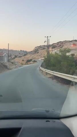 The video captures a view from inside a vehicle driving along a winding paved road. The scene is a rural, hilly landscape with sparse vegetation and rocky terrain. The road has a white dashed line marking the center and a guardrail on the right side. Power lines and wooden utility poles are visible along the roadside, stretching across the frame.

In the background, the hillside is dotted with scrubby bushes and rocky outcrops. Some construction or development appears to be taking place on the right side of the road, with visible structures and what looks like excavated earth. A few traffic signs are present, including a warning sign with an exclamation mark and a triangular symbol, and another sign with text that is too blurry to read. Some orange traffic cones are also visible near the road.

The time of day appears to be late afternoon or early evening, as indicated by the soft, warm light and the clear, pale sky. The weather seems to be calm and fair. The overall mood is peaceful and serene, characteristic of a scenic drive through a rural area. The location is Armenochori Community, Cyprus, which suggests a Mediterranean setting. There are no people visible in the frame.