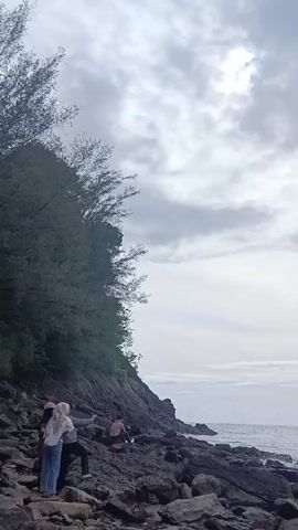 Here's a description of the video:
The video shows a coastal scene on a cloudy day. In the foreground, there is a rocky shore. Several people are present: one group of three individuals, two of whom wear headscarves and are taking photos, and another person sitting a short distance away. The rocks extend to the edge of the sea, where waves break against the shore. To the left, a lush, green hillside covered in trees slopes down to the rocky beach. The sky is overcast with gray clouds, and a diffused sun is barely visible through the clouds. The overall scene conveys a sense of peacefulness despite the cloudy weather.