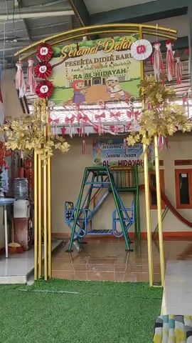 A short video clip shows the entrance to a building, decorated with a welcome banner and festive streamers. The banner reads "Selamat Datang Peserta Didik Baru" and features cartoon images of children playing. To the left of the entrance, a blue swing set is visible, and a water dispenser is positioned near a brown wooden door. The ground is covered with artificial turf, and a white table draped with a black cloth is set up in front of the building. The overall impression is a welcoming setup for a school or educational institution.