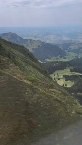 The video shows a slow drone shot descending over a lush green mountainside. The camera pans down and to the right, revealing a valley below dotted with trees and fields. In the distance, more rolling hills and a hazy sky are visible. There are no people or discernible objects in the video, only the natural landscape.