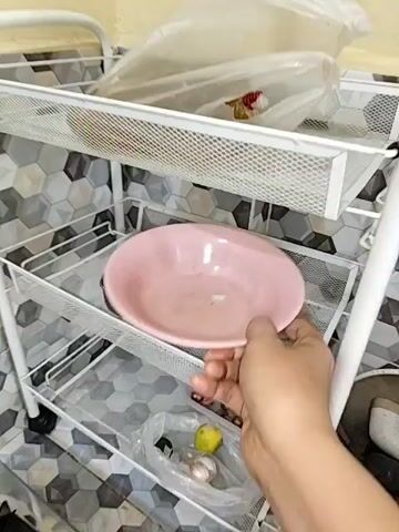 A hand is holding a pink bowl and places it into a kitchen sink. The sink is made of stainless steel and has a faucet and a drain. There is a white dish rack with dishes in the background. The walls are tiled with a hexagonal pattern.