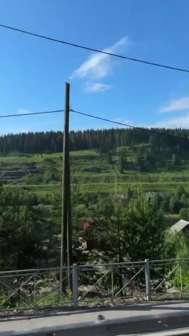 The video shows a landscape with rolling green hills under a clear blue sky. In the foreground, there's a metal railing, possibly part of a bridge or overpass. Beyond that, a small building with a red roof is visible, situated near a body of water. Trees, both dense and sparse, dot the landscape, with a line of tall trees forming a backdrop along the crest of the furthest hill. The overall scene suggests a rural or semi-rural environment on a sunny day.