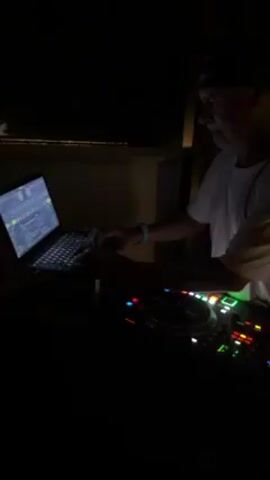 The video shows a DJ operating equipment in a dimly lit environment. The focus is on a laptop displaying music software with waveforms and a track list, alongside a DJ controller with colorful lights and knobs. An adult is seen adjusting the controller while the music plays, their face partially visible. The setting suggests a nightclub or DJ booth.