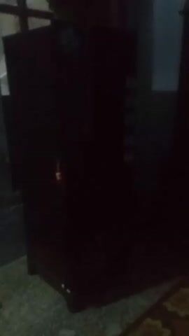 The video shows a dark brown refrigerator standing in a room. The room is dimly lit, and the floor appears to be tiled. There's a window to the left of the refrigerator. The refrigerator has a label on top and several compartments visible on its side. The camera appears to be handheld and slightly shaky.