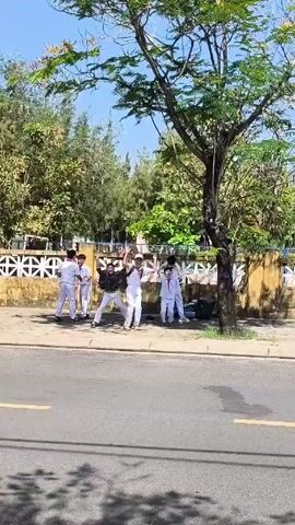 The video shows a group of young people in white uniforms, likely students, standing on a sidewalk next to a road on a sunny day. Most of them are wearing white shirts and pants, with one individual in the center wearing a black jacket and white pants. They appear to be celebrating, jumping, and raising their hands in the air. Behind them is a wall with a decorative pattern, trees, and other greenery. A tree stands between the group and the camera. The road has a yellow line marking a lane, and there are shadows cast by the tree on the road.
