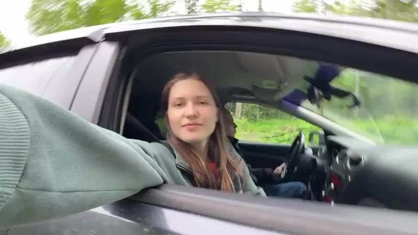 A young woman in a car smiles, leans into the open driver-side window, and waves a hand. The car travels along a road bordered by green trees. The driver is partially visible.
