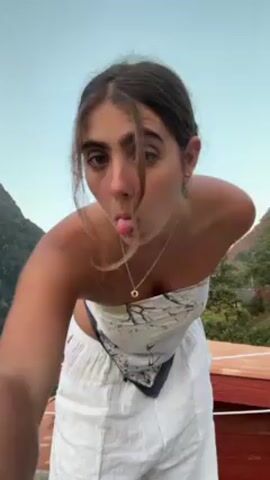 A young woman stands on a wooden deck outdoors, with a mountain range visible in the background. She is wearing a white and blue printed scarf as a top, along with white wide-legged pants. She pulls strands of hair to the front of her face and makes a playful expression at the camera.