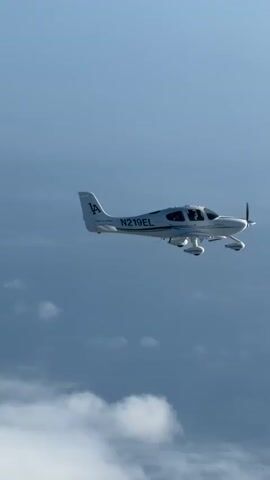 A single-engine airplane, identified by the registration number N219EL and the letters "LA" on its tail, flies through a cloudy sky. The aircraft is white with black accents and has two visible occupants in the cockpit. The camera appears to be positioned slightly below and to the side of the airplane, capturing it in flight against a backdrop of blue sky and large, white clouds. The propeller is in motion, indicating the plane is actively flying.