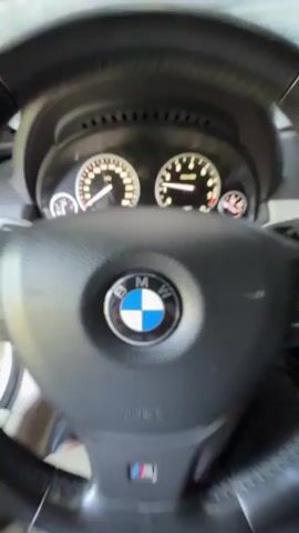 This media file captures the interior of a BMW vehicle, focusing on the dashboard and steering wheel. The scene is brightly lit, suggesting it's daytime with good weather, possibly sunny given the strong illumination. The setting appears to be outdoors, perhaps in a driveway or a yard, indicated by the glimpse of greenery and foliage visible through the windshield, and what looks like a shaded area cast by sunlight.

The primary objects are the car's interior elements: a leather-wrapped steering wheel with the BMW logo prominently displayed in the center, an "M" badge on the lower spoke, and various control buttons. Behind the steering wheel are the car's gauges, showing what appears to be speed and RPM readings. To the right, a part of the infotainment system and climate controls are visible. The presence of the "AIRBAG" text on the steering wheel is also a notable detail.

The activity taking place is the car starting up or being engaged, evidenced by the slight blur and movement in some frames, and the illuminated dashboard. There are no people clearly visible, but the perspective suggests someone is seated in the driver's seat. The overall mood is functional and neutral, focusing on the car's operational status.

While the specific location is Novy, Russia, the visual cues are more general to a car interior and an outdoor environment. No specific emotional context is conveyed. Text visible includes "BMW", "AIRBAG", and potentially "M" on the steering wheel, as well as the numbers and indicators on the car's dashboard.