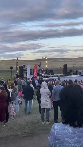 A crowded outdoor concert event is taking place. A stage with lighting and speakers is set up in the middle ground. People are gathered in front of the stage, some standing and some sitting on the ground. The environment appears to be a grassy, hilly area with a cloudy sky. Several people are visible in the foreground, with one person in a patterned blanket looking towards the stage. Another woman in a light-colored jacket is holding a cup and looking to the right. In the distance, more people are spread out on the hills. A kite is visible flying in the sky.