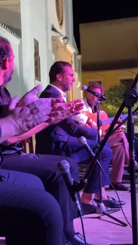A medium shot shows three men seated on a wooden stage performing music outdoors at night. The man in the foreground on the left is clapping his hands. He is wearing a black shirt. The man in the center, wearing a dark suit jacket and white shirt, is singing into a microphone and clapping his hands. The man on the right, wearing a light-colored shirt and dark pants, is playing an acoustic guitar. Another guitar and microphone are visible on the stage. The background shows a white building with a large arched window and a tree. The lighting is purple and warm yellow, creating a dramatic atmosphere.