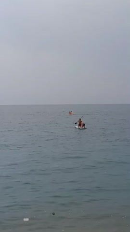 The video shows two people on a paddleboard in the ocean. There is another person on a different paddleboard further in the distance. The sky is overcast and the water is calm.