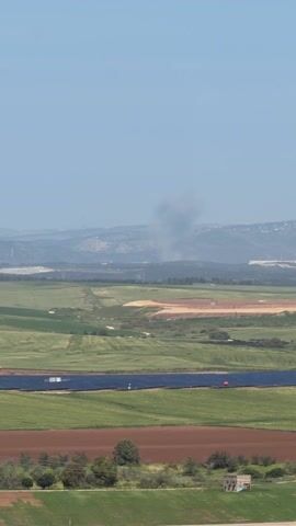 The video shows a distant landscape under a clear, blue sky. Green and brown fields stretch into the distance. A solar panel array is visible in the foreground. A plume of dark smoke rises from the distant hills. The smoke appears to be emanating from a specific location.