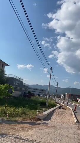 The video shows a street scene on a sunny day. In the foreground, there is a dirt path with some debris. To the left, there is a building with the words "RADANOVIĆ GARAGE" visible. There are power lines stretching across the frame, supported by utility poles. In the background, there are more buildings and a mountain range under a blue sky with scattered clouds. The road in the middle ground appears to be under construction or newly paved. A red tractor is parked near the garage.