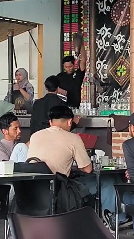A group of people are gathered at what appears to be an outdoor cafe or food stall. In the background, a man in a black t-shirt is holding up a broom or similar object, seemingly demonstrating something or preparing for a task. A woman wearing a hijab is standing nearby, looking in the same direction. Several other individuals are seated at tables in the foreground, with one man in a light brown shirt facing away from the camera. The environment suggests a casual gathering, possibly in a Southeast Asian setting, with decorative banners and a wooden structure visible.