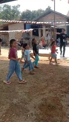A group of children are participating in a game outdoors, possibly a race or a relay. They are lined up, and some are running or moving forward. One adult male in the background appears to be directing or observing the children. The setting is a yard with a building in the background, decorated with red and white banners, suggesting a celebration or national event. The ground is covered with dirt and grass. Other people are seated or standing on the sides, watching the children play. The overall atmosphere seems festive and informal.