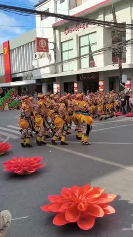The video shows a street parade of performers in colorful costumes. The performers are in tight formation, with most wearing yellow, orange, and black outfits with headdresses. They are moving forward in unison, with one performer in front wearing a more elaborate costume of green, red, and yellow, dancing and leading the group. The parade is taking place on a city street lined with buildings. There are flower decorations on the ground, and a crowd of spectators is visible behind barriers on the sides of the street. The sky is partly cloudy. Businesses such as Chowking, McDonalds and Grace Pharmacy are visible in the background.