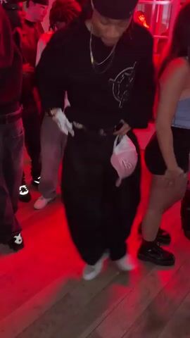Here is a detailed description of the video:
The video shows a group of people dancing in a room with a red light atmosphere. The focus is on a person wearing a black beanie, a black sweatshirt with a white design, loose black pants, and white sneakers. They are holding a white plastic bag in one hand and dancing, with their arms moving and body swaying to the music. Other people are around them, also dancing or standing by. Some details are visible, such as a fur coat, jeans, boots, and a door with white frames. The floor is made of wood panels.