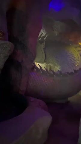 The video shows a statue of Ganesh sitting cross-legged on a platform. The statue is golden and adorned with skulls around its base. The statue is illuminated by purple and blue lights, creating a mystical atmosphere. The background appears to be a cave or rock formation, with rough textured walls. The video pans slightly, showing different angles of the statue and its surroundings. There are no people visible in the video, and no actions or interactions are depicted beyond the panning camera movement.