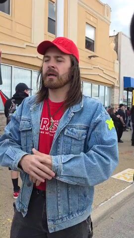 The video shows a man with long brown hair and a beard standing on a sidewalk. He is wearing a red baseball cap, a red T-shirt, a jean jacket with a yellow star on the sleeve, and gray pants. He is standing with his hands clasped in front of him, and he is speaking. Behind him, there are several people standing and holding a flag with green, white, and black stripes. The background is a building with a yellow facade and windows. The sidewalk is made of concrete and there is a curb separating the sidewalk from the street. The scene appears to be a protest or demonstration.