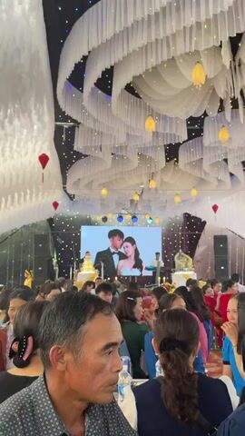 The video captures an indoor wedding reception in Bình An, Vietnam, with numerous guests seated at tables. A large screen at the front of the room displays a rotating series of professional photographs of a young couple, presumably the bride and groom, dressed in wedding attire. On either side of the screen, there are elaborate, multi-tiered wedding cakes, and what appears to be a live band or musical ensemble is set up, including a keyboard player and a drummer.

The ceiling is elaborately decorated with white draped fabric, creating a wave-like pattern, and interspersed with hanging lanterns emitting a warm glow. Stage lights illuminate the area around the screen. Guests are casually dressed, seated, and observing the festivities, with some talking amongst themselves. The atmosphere appears lively and celebratory. No specific text is clearly visible beyond the images on the screen.