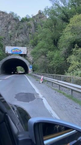 The video shows a car driving into a tunnel. The tunnel entrance is concrete and has a large sign above it with graffiti. The road has white lane markings and there are red and white striped posts on the side. The car's side mirror is visible in the foreground. Inside the tunnel, the road is paved and lit by overhead lights and the headlights of other vehicles. The tunnel walls are grey and textured. As the car progresses, the exit of the tunnel becomes visible as a bright circle of light.