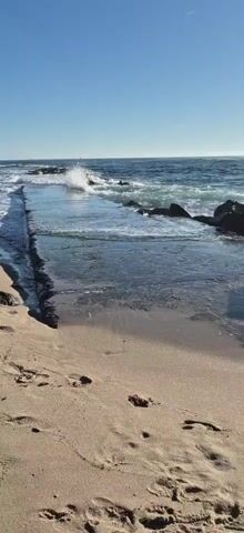 The video shows a beach with footprints in the sand. Waves are crashing on rocks in the ocean. The sky is clear and blue.