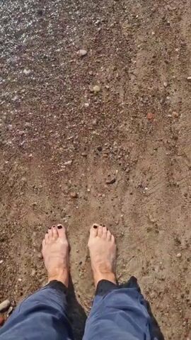 The video shows a pair of bare feet with black toenail polish standing on a sandy beach. The waves are gently washing over the feet and the shoreline. The water is clear and the sand is a mix of brown and grey pebbles. In the distance, the ocean stretches out to the horizon under a cloudy sky.