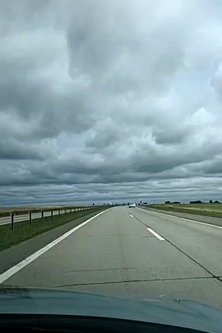 A first-person perspective video shows a highway on a cloudy day. The road is asphalt with white lines dividing the lanes. There are guardrails and grassy fields on the left side of the road. Several vehicles, including a white truck and a few cars, are visible in the distance on the highway. The sky is filled with heavy, dark gray clouds, suggesting overcast weather. The footage appears to be captured from inside a moving vehicle, as indicated by the dashboard visible at the bottom of the frame.