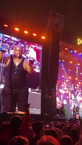The video shows a concert crowd in front of a large stage. A man in a black tank top and shorts is on the left side of the stage, looking towards the audience. Behind him, a large screen displays a colorful, abstract image. Above the stage, the words "PARK LIVE" are illuminated in large yellow letters. The stage is lit with purple and red lights, and there is smoke or haze in the air. The crowd is mostly visible from behind, with many people holding up their phones to record the performance. The overall atmosphere is energetic and festive.