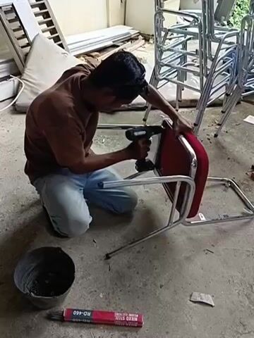 A man is kneeling on the floor, assembling a red chair. He is wearing a brown t-shirt and blue jeans, with safety glasses on his forehead. He holds a cordless drill in his right hand and is using it to screw into the metal frame of the chair. The chair has a red cushioned seat and back. Next to the man is a metal bucket filled with screws and a box of welding rods labeled "NIKKO STEEL RD-460". In the background, there are stacks of similar chairs, and some building materials, including what appears to be window frames and wooden planks. The setting looks like a workshop or storage area, possibly in an outdoor or semi-outdoor space given the presence of natural light and the slightly dusty floor. The overall atmosphere appears to be one of focused work. The time of day is not explicitly indicated, but the lighting suggests it could be daytime.