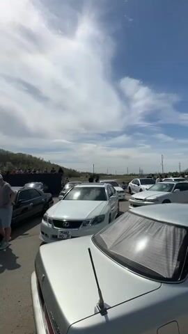 The video shows a crowd of people gathered around several cars parked in an outdoor lot. Most of the cars are various models of Toyota and Lexus, with one red sports car also visible. Some of the cars have their hoods open, with people looking inside the engines. There is a slight hill with some trees and shrubbery behind the cars. The sky is mostly blue with white clouds, and the sun is shining.