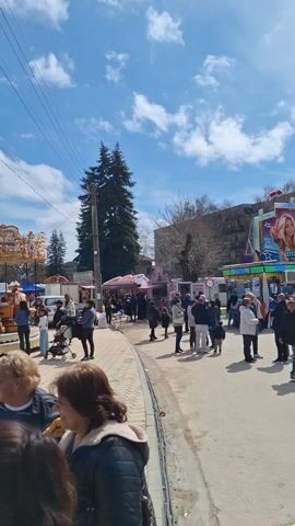 Here's a concise description of the video:

The video shows a lively fairground scene during the daytime. Many people, including adults and children, are walking around and enjoying the attractions. There are several fair rides including a "Summer Games" attraction featuring images of young women, and a Ferris wheel. Food stalls selling sweet corn are also visible. The weather is pleasant with a blue sky and scattered clouds. The area appears to be paved and surrounded by buildings and trees.