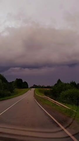 The video shows a road leading into the distance under a heavy, dark sky. The road is paved and has a white line marking the lanes. There is a guardrail on the right side of the road. Trees line the left side of the road, and fields of vegetation are visible on both sides. A few cars are seen on the road in the distance. The sky is filled with dark, ominous clouds, suggesting an approaching storm. The overall mood is one of a journey through a dramatic natural landscape.