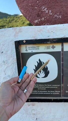 A hand holds a joint and a lighter in front of a sign that depicts a crossed-out fire symbol. The sign also contains text in Catalan and Spanish, indicating it is from the Generalitat Valenciana. The background shows a red, textured object and a hillside. The person's hand has a ring on one finger. The overall scene appears to be outdoors.