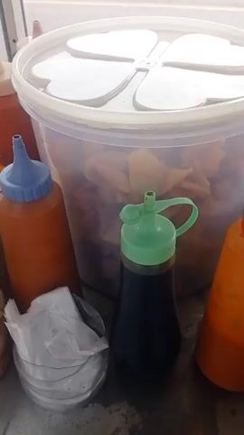 The video shows a close-up view of a food cart, likely selling meatballs and other fried items. In the foreground, a white metal frame with orange circular logos is visible, partially obscuring the view. To the left, an orange squeeze bottle with a red cap sits next to a clear plastic container filled with fried food. The main focus of the video is a stainless steel tray divided into compartments. These compartments are filled with numerous small, round, light brown meatballs. One compartment contains pieces of golden-brown fried food, possibly tofu or tempura. The background is blurry but appears to be an outdoor setting with a paved surface. The lighting suggests it is daytime.