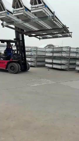 A forklift is moving large, angular panels. The forklift, which is red and black and says “A50” on the side, is driven by a man wearing blue clothing. The forklift is carrying three large white and silver panels on a metal frame. The panels are pointed at the top. The forklift is driving on a concrete surface in a large outdoor area. Stacks of similar panels are visible in the background, wrapped in clear plastic and held together with metal framing. The weather appears overcast.
