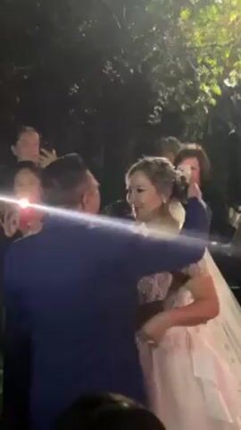 The video shows a bride and groom embracing during an outdoor wedding reception. The bride wears a white, sleeveless gown with floral embroidery, and a bracelet on her left wrist. She holds a bouquet of white flowers. The groom is in a dark blue suit. They embrace tightly in the center of the frame. 
In the background, people are taking photos and videos of the couple with their phones. A person in a navy dress holds up a light source, possibly a phone flashlight. The event appears to be taking place at night, with dark surroundings and lush greenery visible in the background.