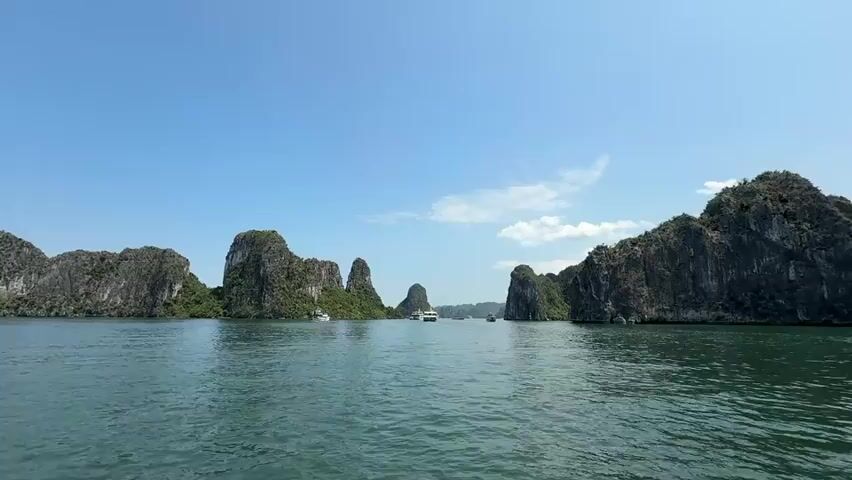 The video captures a wide shot of a serene bay on a bright, sunny day. Several rocky, island-like land formations dot the scene, covered with green vegetation. The calm water reflects the sky, which is a clear blue with some wispy clouds. There are a few boats navigating the water between the islands. The overall scene conveys a sense of peacefulness and natural beauty.