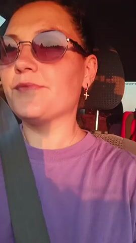 A woman with dark hair pulled back is wearing sunglasses with pink lenses and a purple t-shirt. She is in the driver's seat of a car, with a seatbelt across her chest. She smiles at the camera as the sun sets, casting a warm glow on her face and the interior of the car.