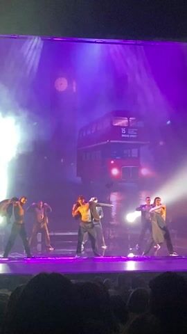The video shows a dance performance on a stage. There are six dancers, all male, arranged across the stage. They are dressed in dark pants and some are wearing open jackets or have removed their shirts. The backdrop of the stage features a large image of a red double-decker bus and what appears to be the Big Ben clock tower in the background. The stage is illuminated with bright purple and white spotlights, and there is a haze or fog effect, adding to the dramatic lighting. The dancers are moving in sync to the music. The perspective is from the audience, indicated by dark shapes along the bottom edge of the frame.
